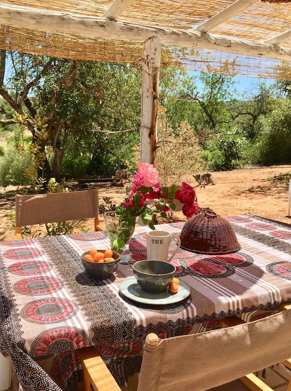 An outdoor dining area with a table set for two, featuring a patterned tablecloth, a woven basket, a vase with flowers, and a bowl of fruit.