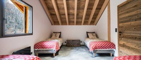 A bedroom with two beds, wooden ceiling beams, a window with a view of trees, and a wooden wall.