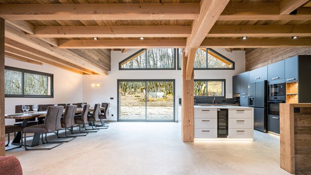 Una cocina moderna con vigas de madera, un comedor con sillas y mesas, y ventanas grandes.
