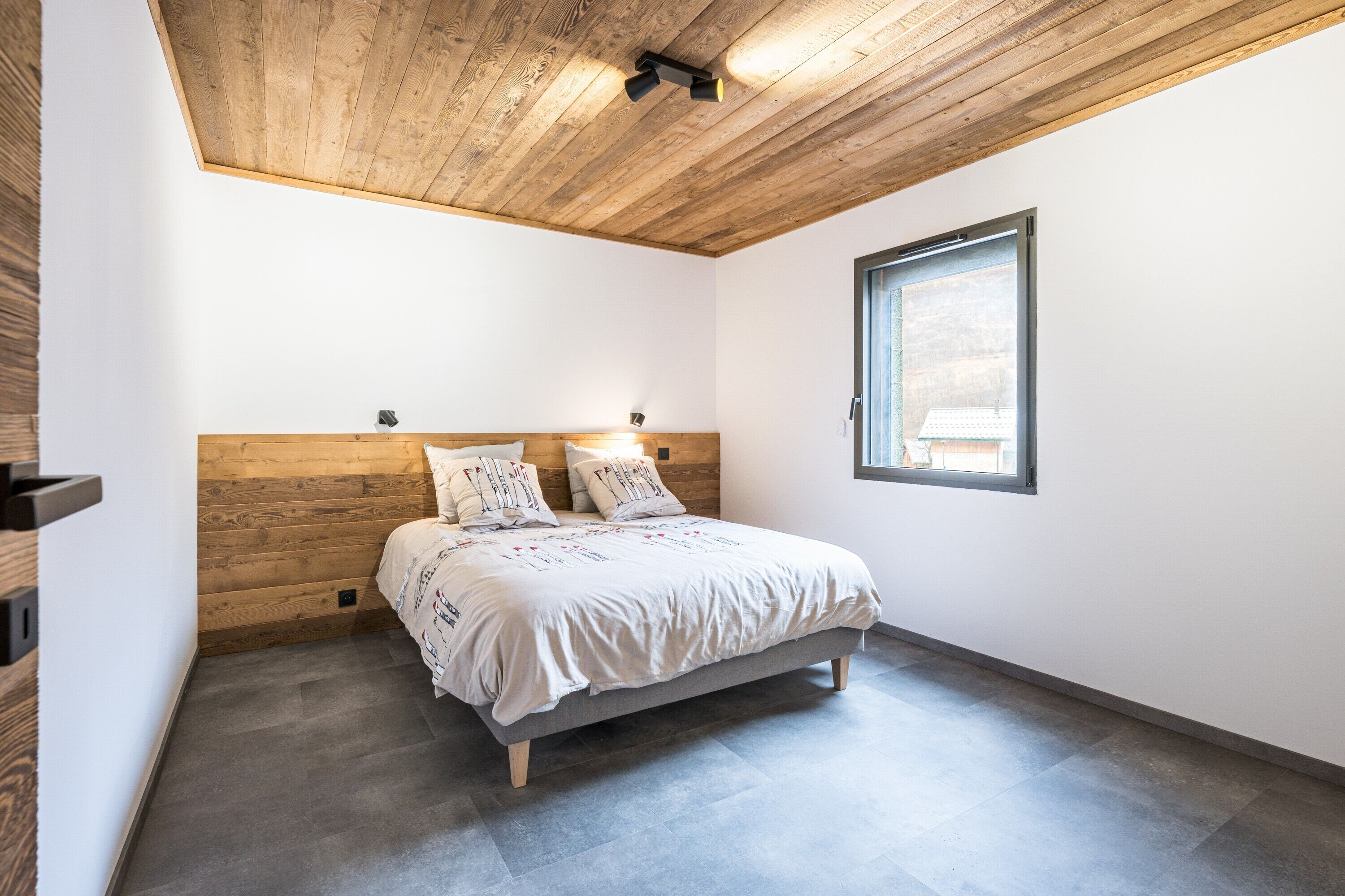 A bedroom with a wooden ceiling, a bed with a headboard, a window, and a wall-mounted light fixture.