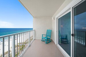 A balcony with a view of the beach and ocean, a turquoise rocking chair, and a glass door.