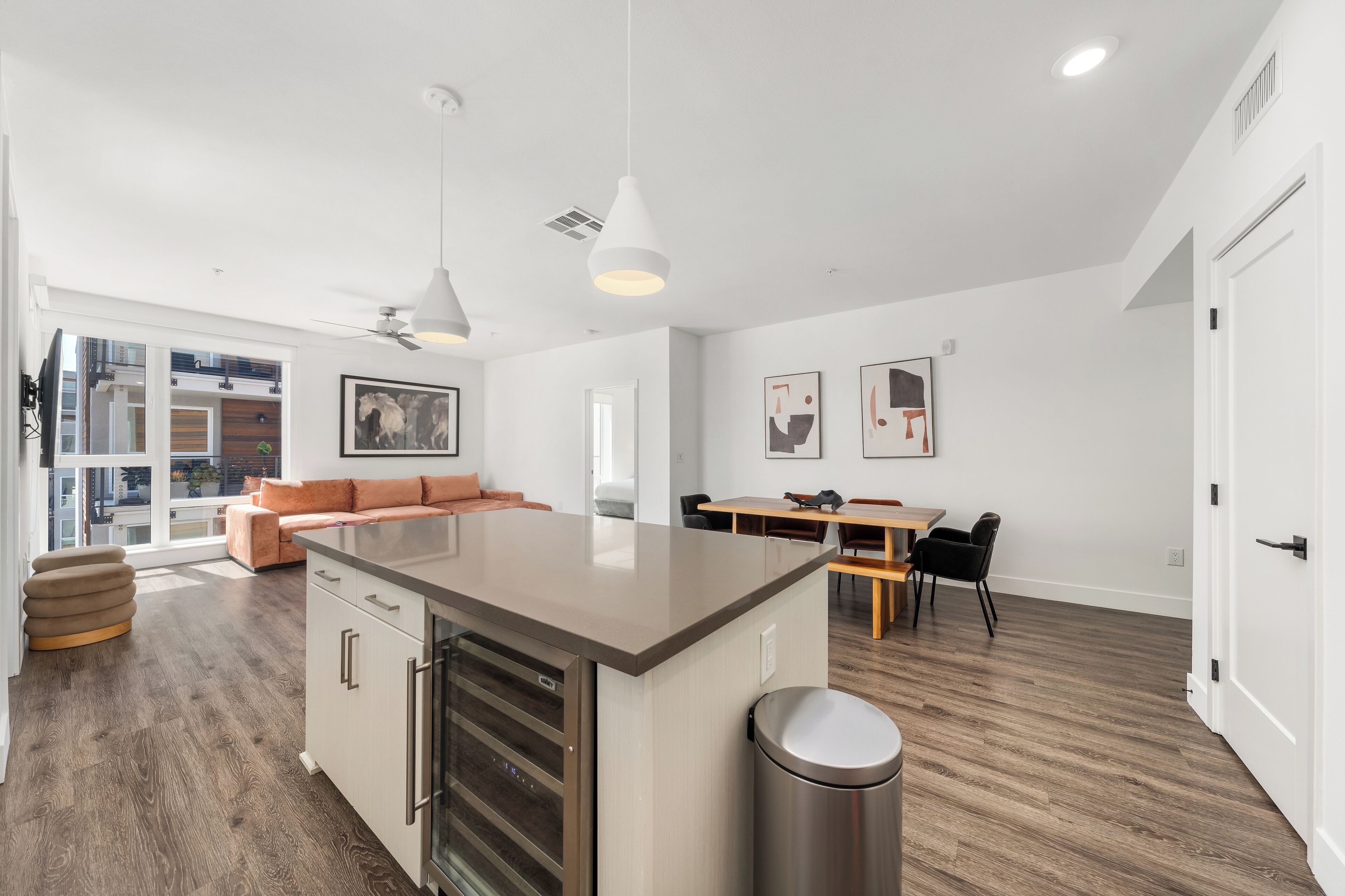 Una cocina moderna con una isla central, una zona de comedor con una mesa y sillas de madera, y una sala de estar con sofá y ventilador de techo.