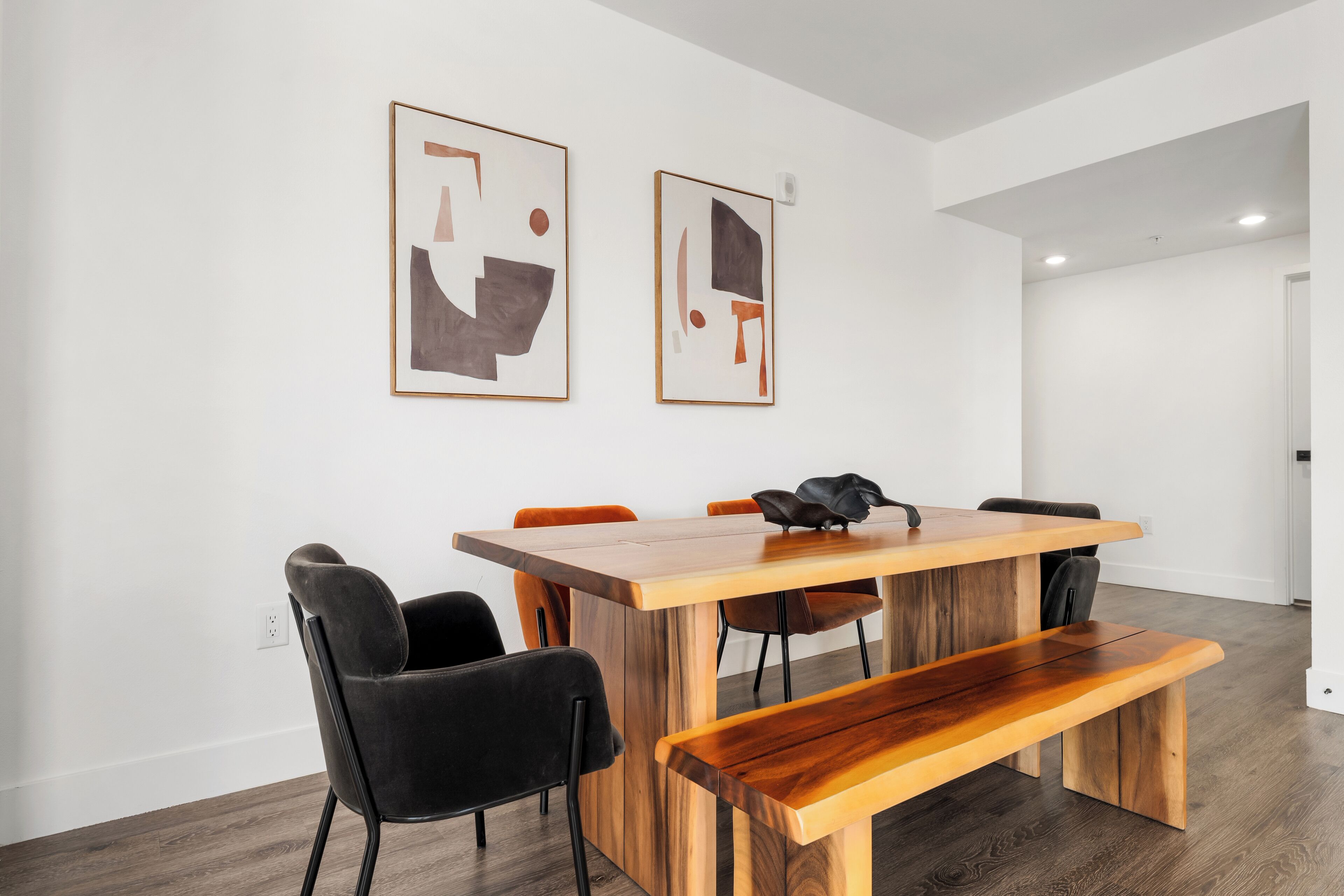 Un comedor moderno con una mesa de madera, sillas negras y un banco. Hay dos cuadros abstractos en la pared.