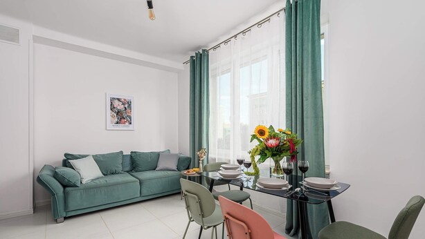 Ein modernes Wohnzimmer mit einem türkisfarbenen Sofa, einem Esstisch mit Stühlen und einem großen Fenster mit grünen Vorhängen.