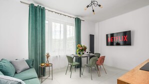 Ein modernes Wohnzimmer mit einem grünen Sofa, einem Esstisch mit Stühlen, einem kleinen Beistelltisch mit Vase und Blumen sowie einem an der Wand montierten Fernseher, der 'NETFLIX' anzeigt.