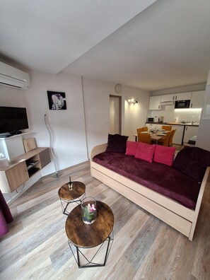 Un salon moderne avec un canapé, une table basse, un téléviseur et, au fond, un coin cuisine.