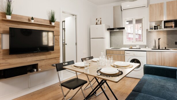 A modern kitchen with a flat-screen TV, wooden cabinets, a built-in oven, and a dining area with a table set for four.
