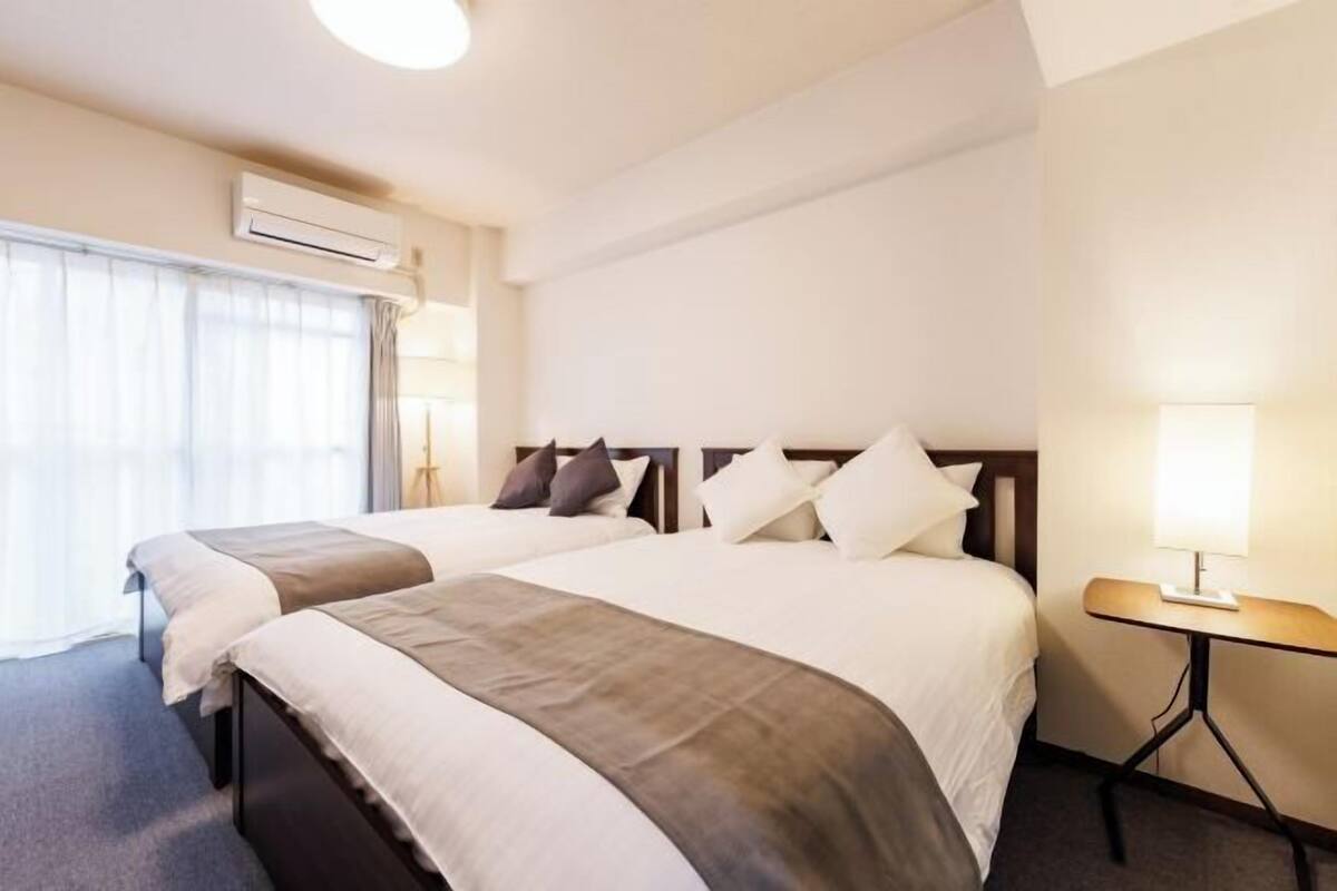 A hotel room with two beds, a bedside table with a lamp, an air conditioning unit, and a window with curtains.