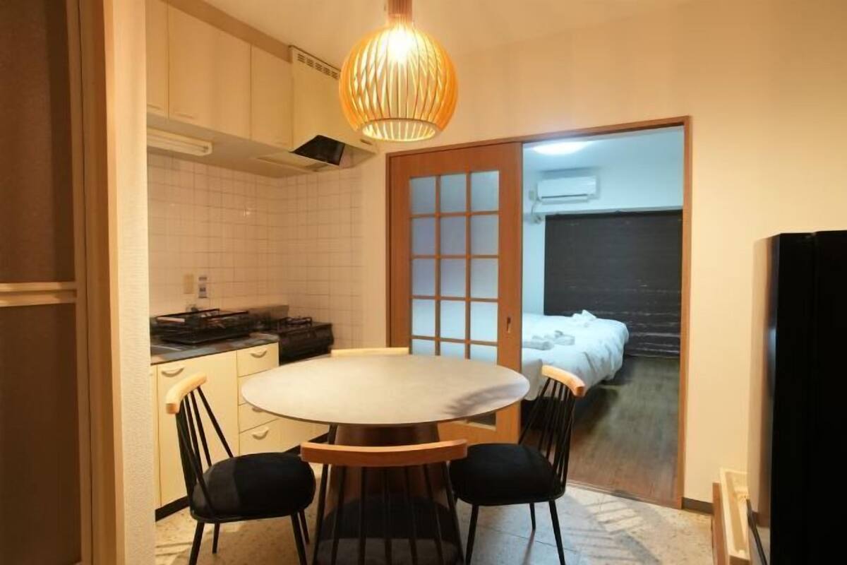 A compact kitchen with a dining table and chairs, a hanging light fixture, and a sliding door leading to a bedroom.