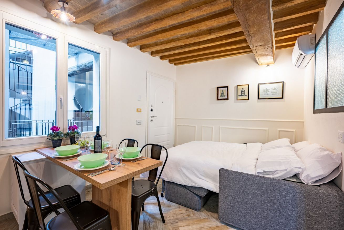 A room with a bed, a dining table with chairs, a window with a view of a spiral staircase, and a ceiling with exposed beams.