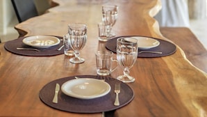 A dining table set with plates, glasses, and cutlery, with a woven pendant light above. - Casinha Praia do Espelho (Porto Seguro)