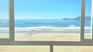 Une vue sur la plage, à travers une fenêtre, avec un ciel dégagé, une mer calme et des montagnes lointaines.