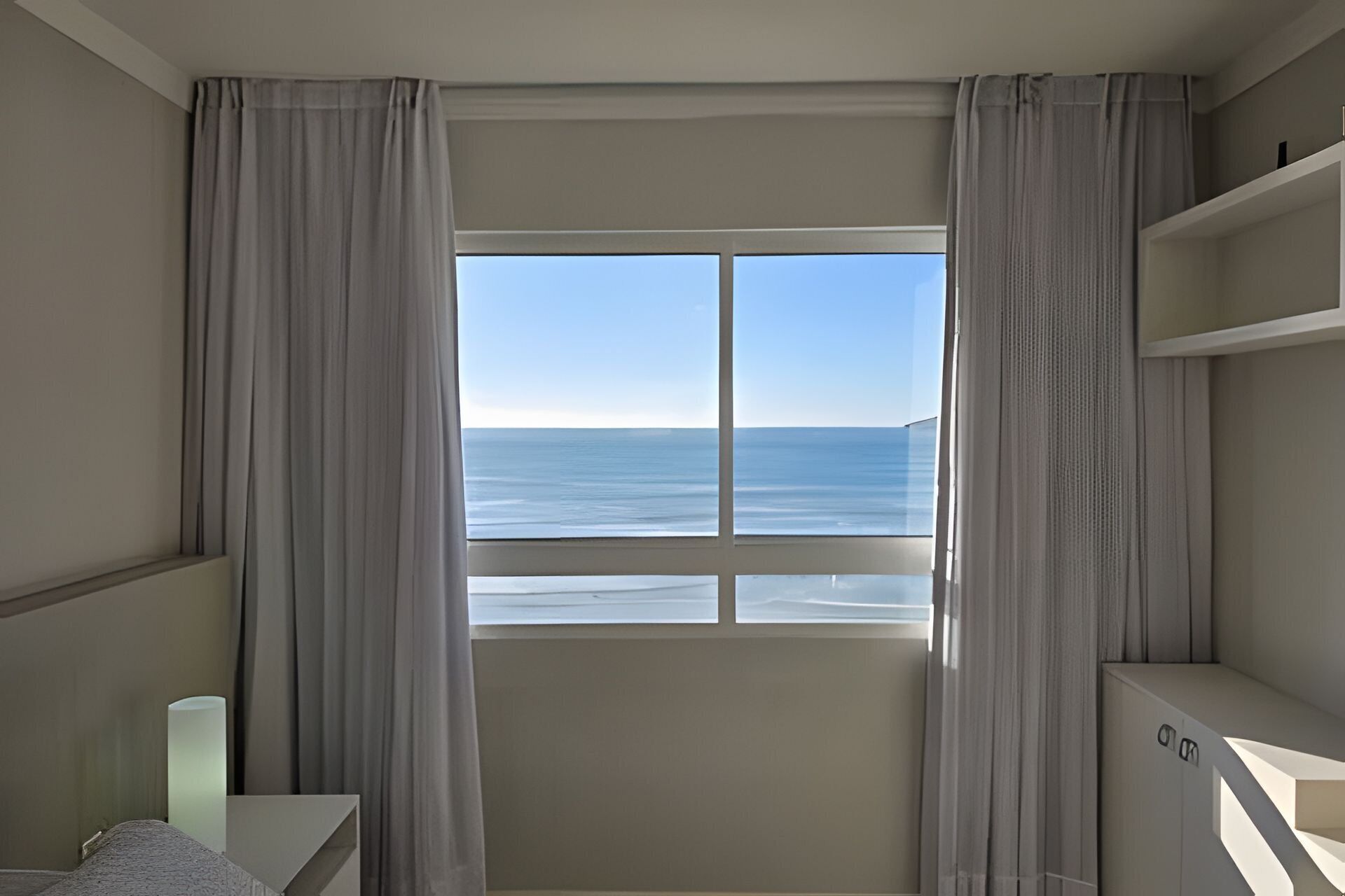 A room with a large window offering a view of the sea. There are curtains on the window, a bedside table, and a small cabinet.