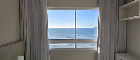 A room with a large window offering a view of the sea. There are curtains on the window, a bedside table, and a small cabinet.