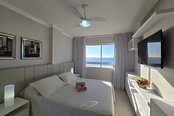Habitación de hotel moderna con cama, ventilador de techo, televisor y ventana con vista al mar.