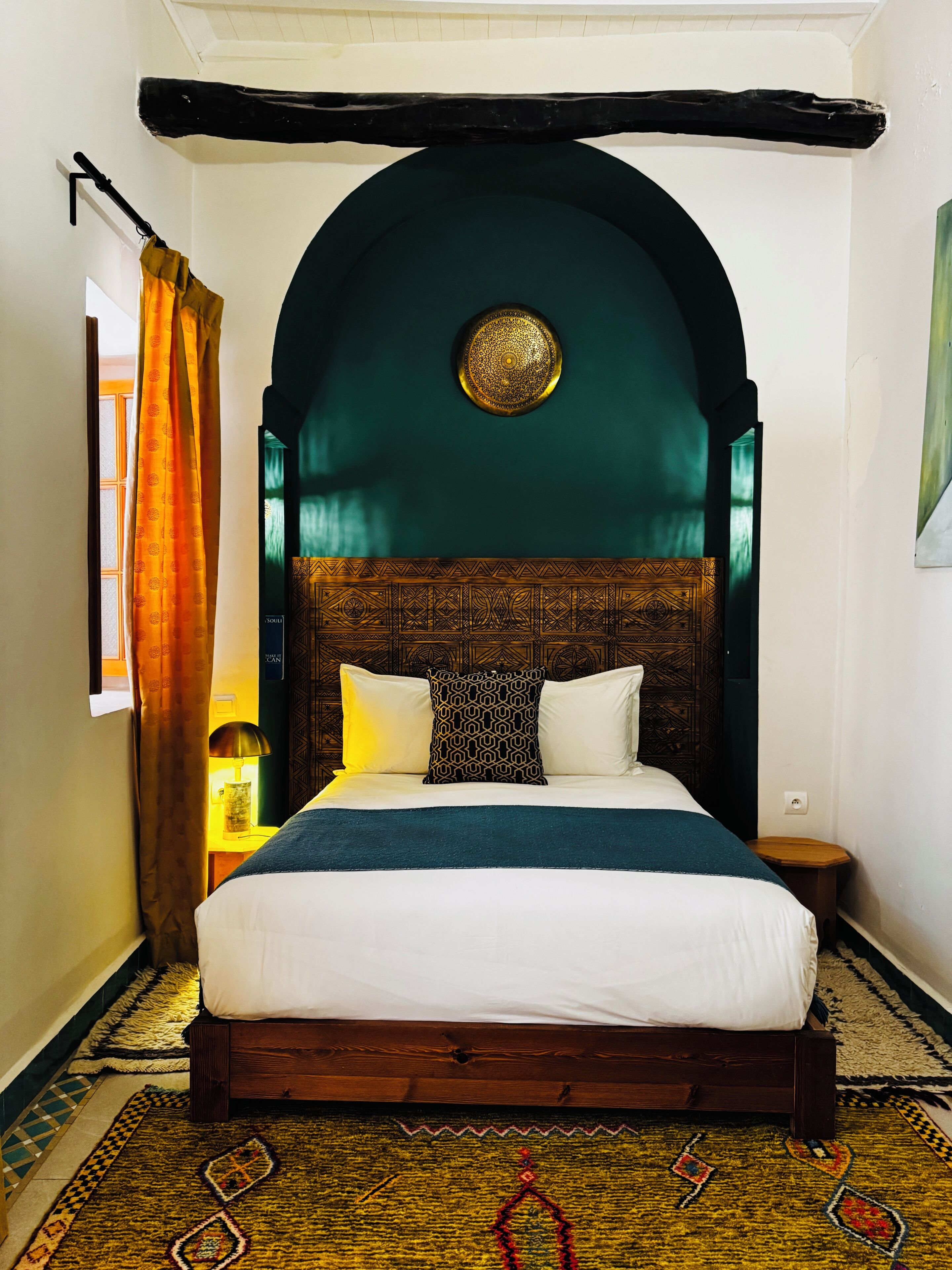 A room with a bed, a patterned rug, a wooden headboard, and a wall-mounted light fixture.