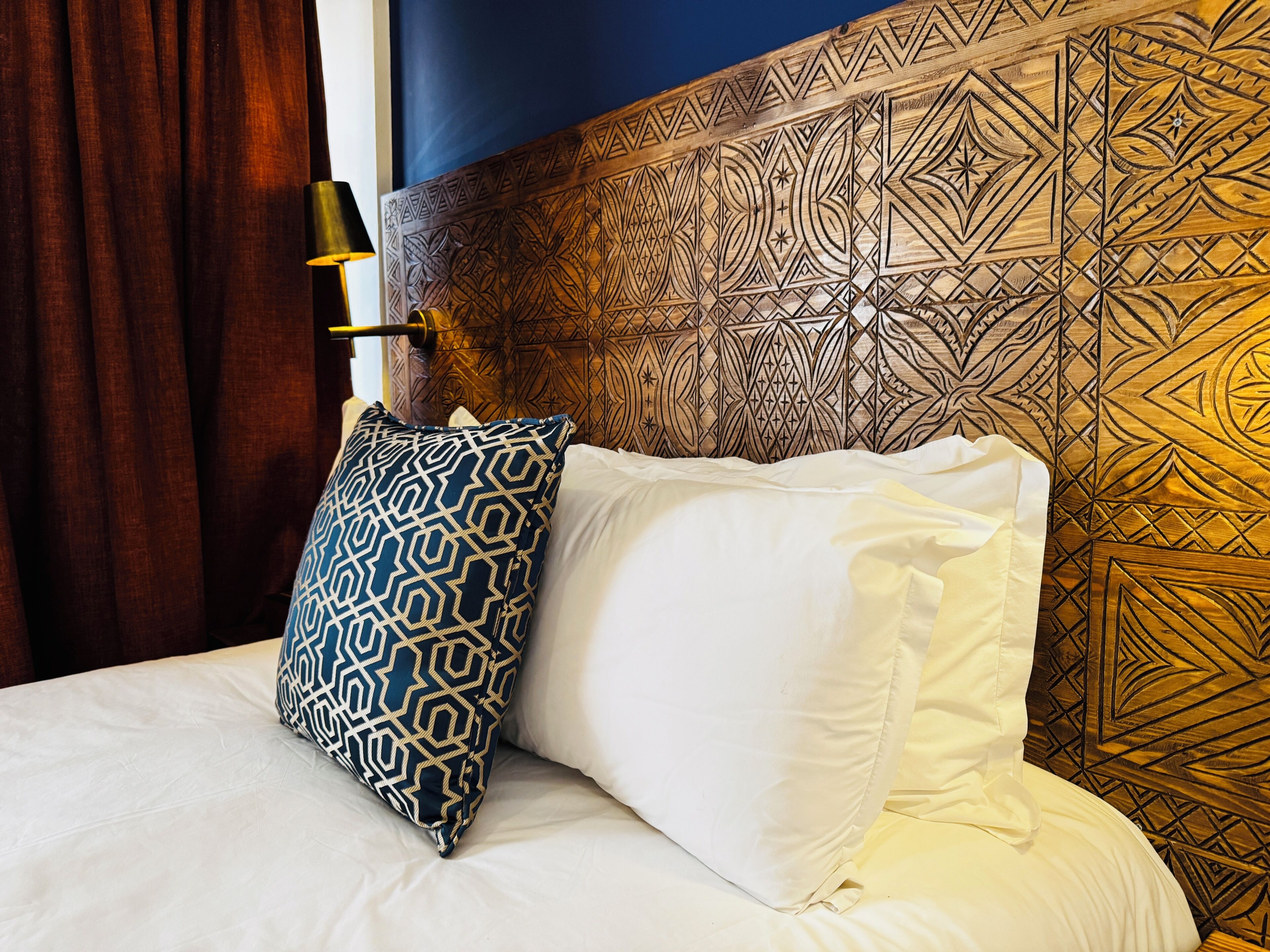 Una cama con almohadas estampadas, un cabecero de diseños elaborados y una mesita de noche con una lámpara.