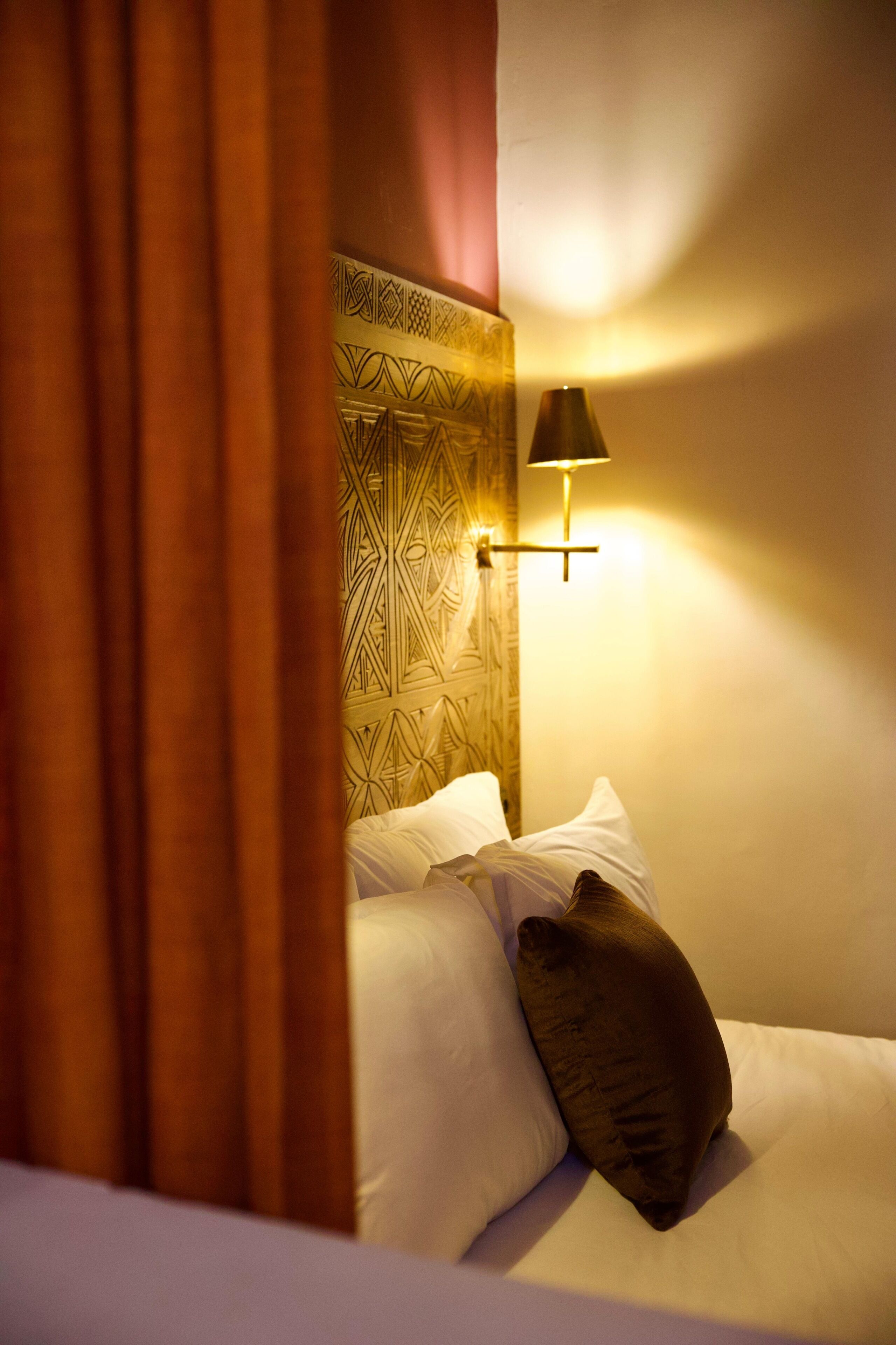 A hotel room with a patterned headboard, a bedside lamp, and a bed with pillows.