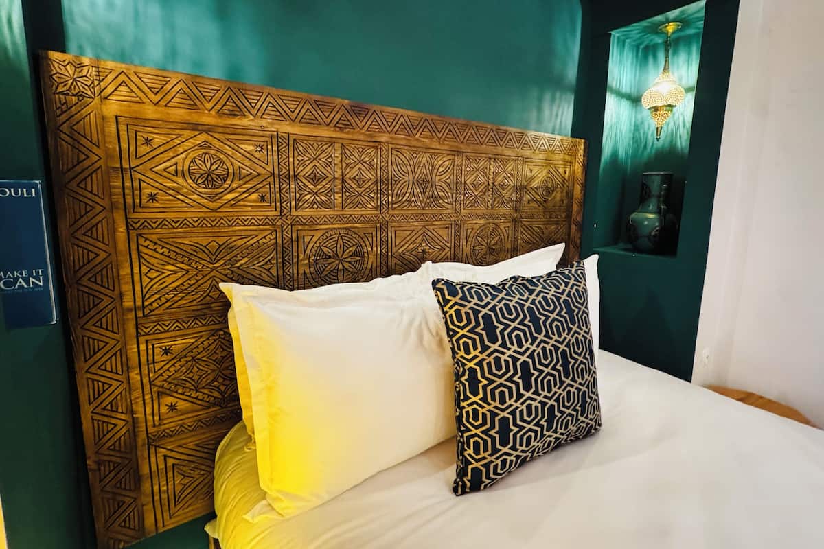 A bed with a patterned headboard, white and patterned pillows, and a wall-mounted light fixture.