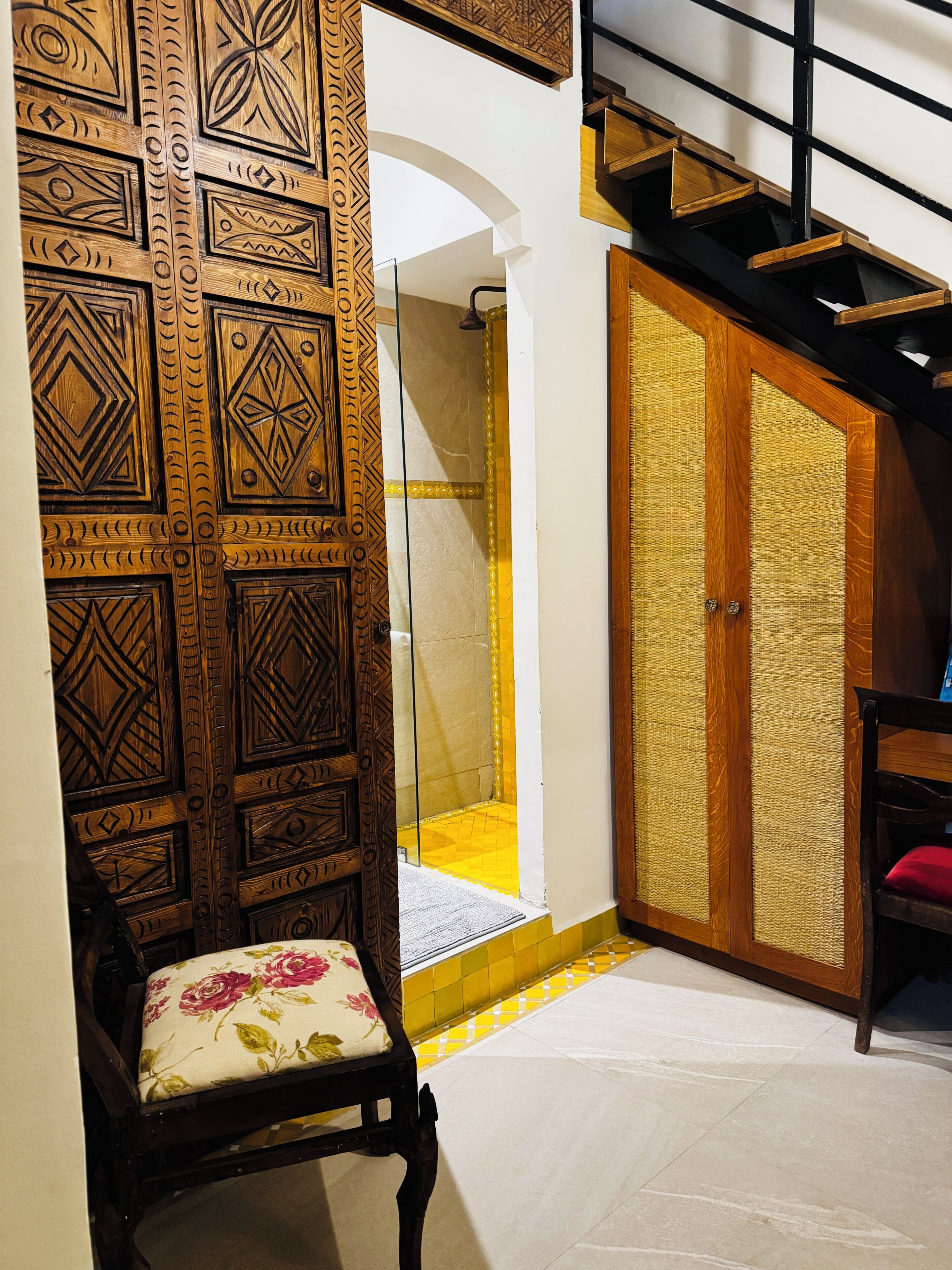 A room with a patterned wooden door, a chair with a floral cushion, a tiled floor, and a staircase with a black railing.