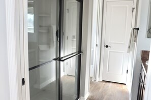Un couloir avec une porte vitrée donnant sur une salle de bain, une porte blanche et un coin cuisine avec des placards.