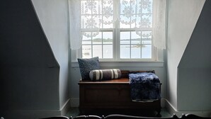 Un dormitorio con techo inclinado, una ventana con cortinas de encaje, un banco de madera con almohadones y una alfombra estampada.