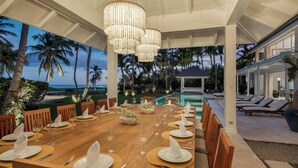 A dining area with a long table set for a meal, overlooking a pool and palm trees.