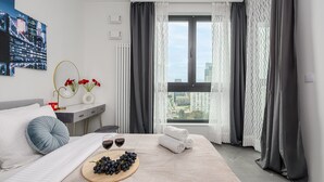 A hotel room with a bed, a bedside table with a vase of flowers, a window with curtains, and a cityscape view. - Lumina 15th Floor Apartment in Warsaw  (Warsaw)