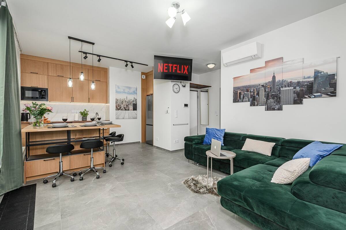 Un salon moderne avec un canapé vert, une petite table sur laquelle se trouve un ordinateur portable, et un coin cuisine doté de meubles en bois et d’un four à micro-ondes.