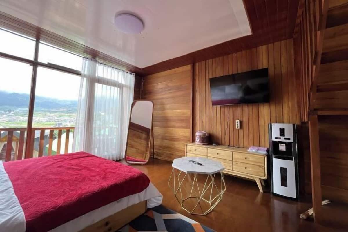 A bedroom with a bed, a TV mounted on the wall, a small table, and a refrigerator.