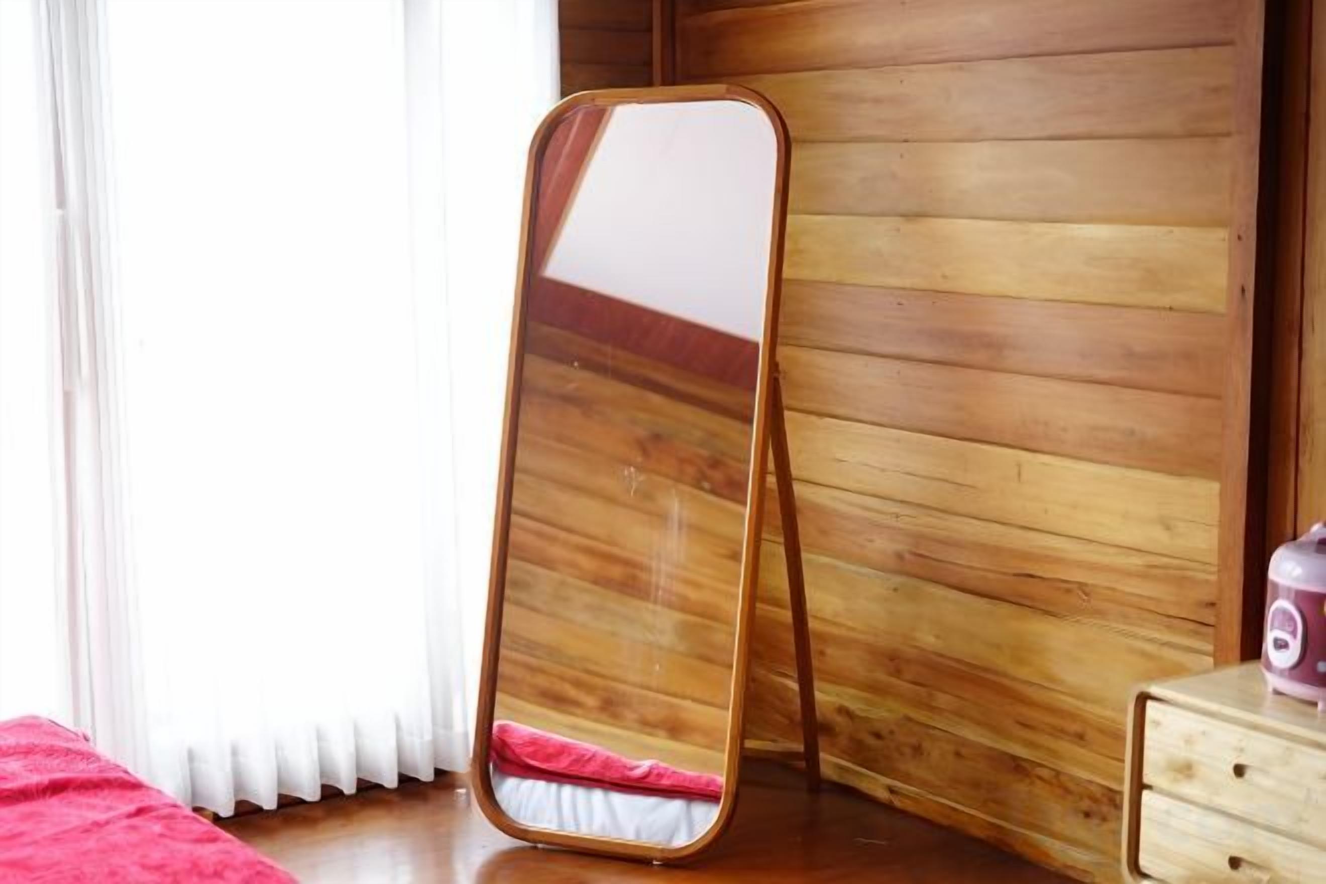 A wooden room with a standing mirror, a wooden dresser, and a window with curtains.