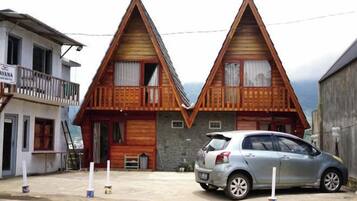 Two wooden houses with a triangular roof design, a car parked in front, and a signboard on the left.