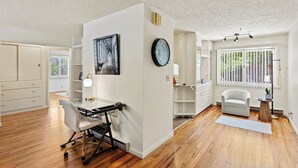 A spacious room with a wooden floor, a desk with a chair, a clock, a chair, and a window with blinds.