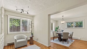 A spacious room with hardwood floors, a dining area with a table and chairs, a gray armchair, and large windows with blinds.