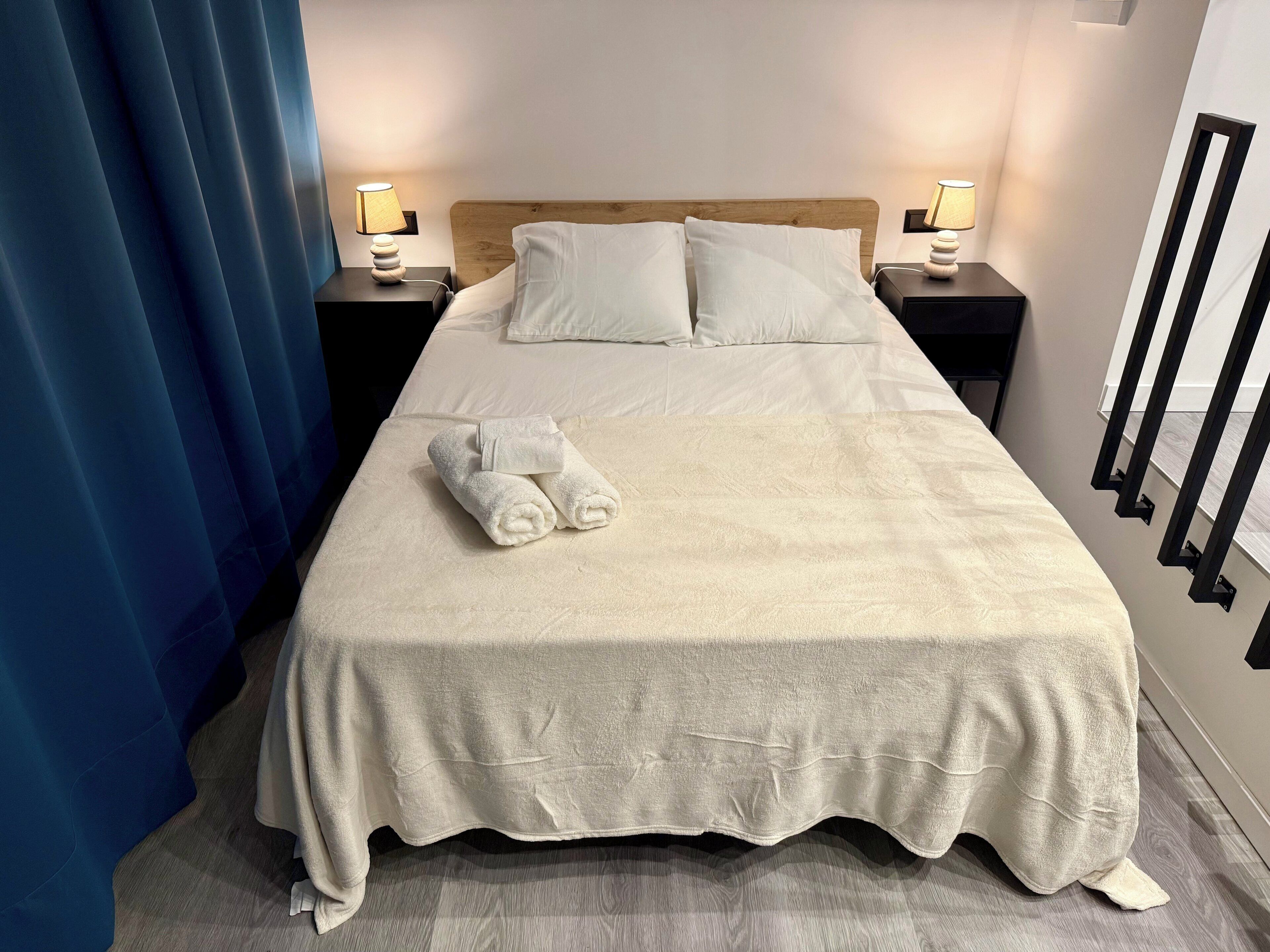 Una cama bien hecha con sábanas blancas, dos mesitas de noche con lámparas y una cortina azul.