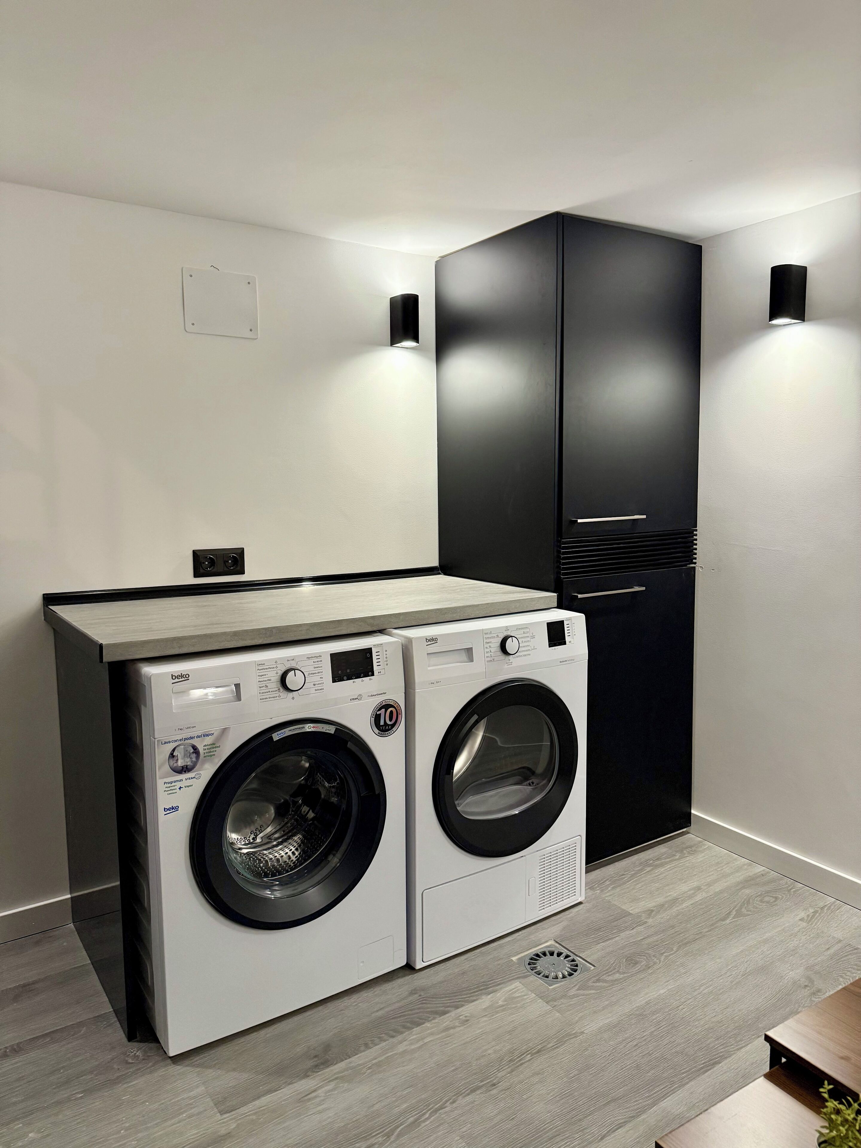 A modern laundry room with two front-loading washing machines, a built-in drying rack, and a black cabinet.