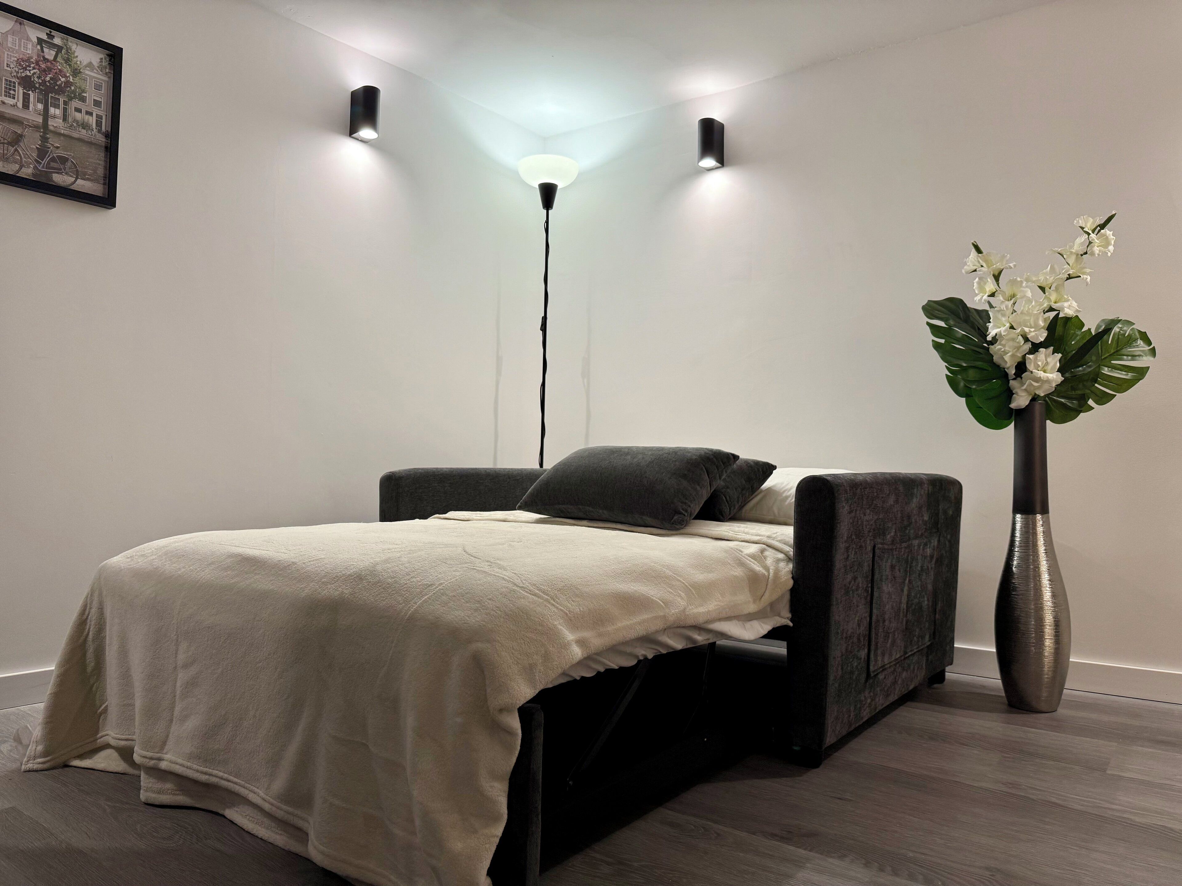 A modern bedroom with a bed, a floor lamp, a vase with flowers, and a framed picture on the wall.