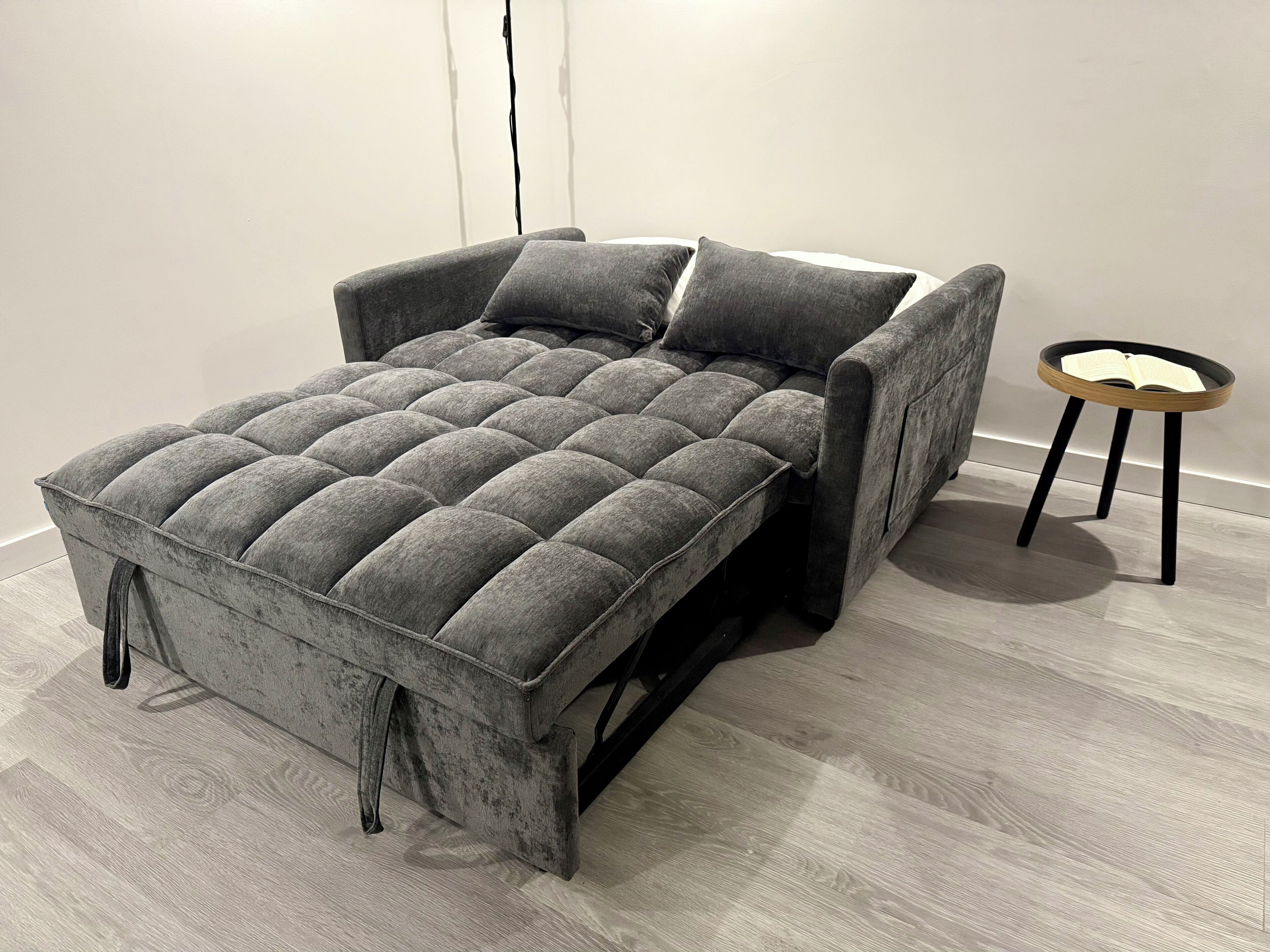 Un canapé moderne, de couleur grise et recouvert de tissu, doté d’un motif capitonné, placé contre un mur uni, avec une petite table d’appoint ronde en bois à côté.