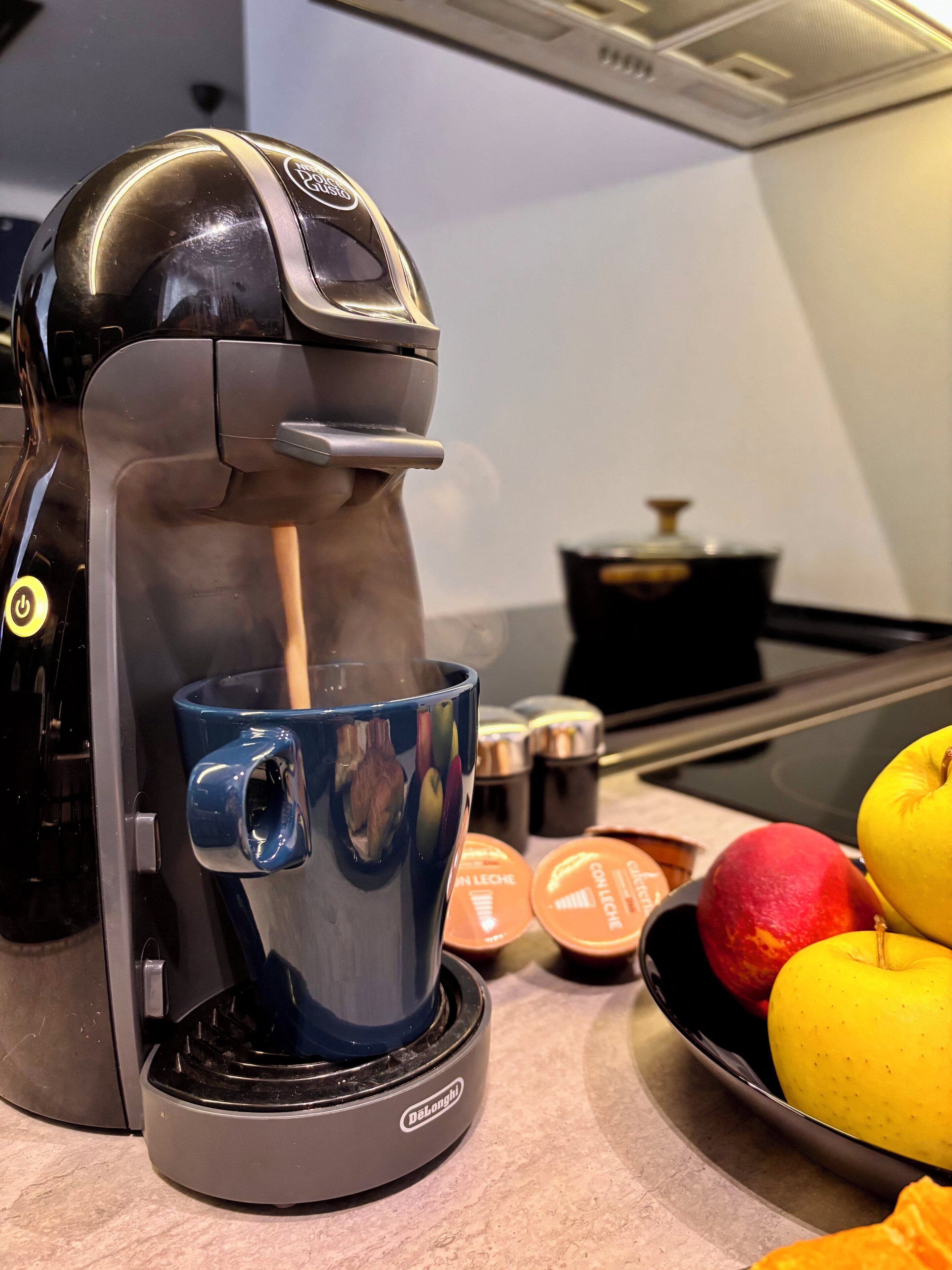A coffee maker with a cup of coffee, a bowl of fruit, and some containers on a kitchen counter.