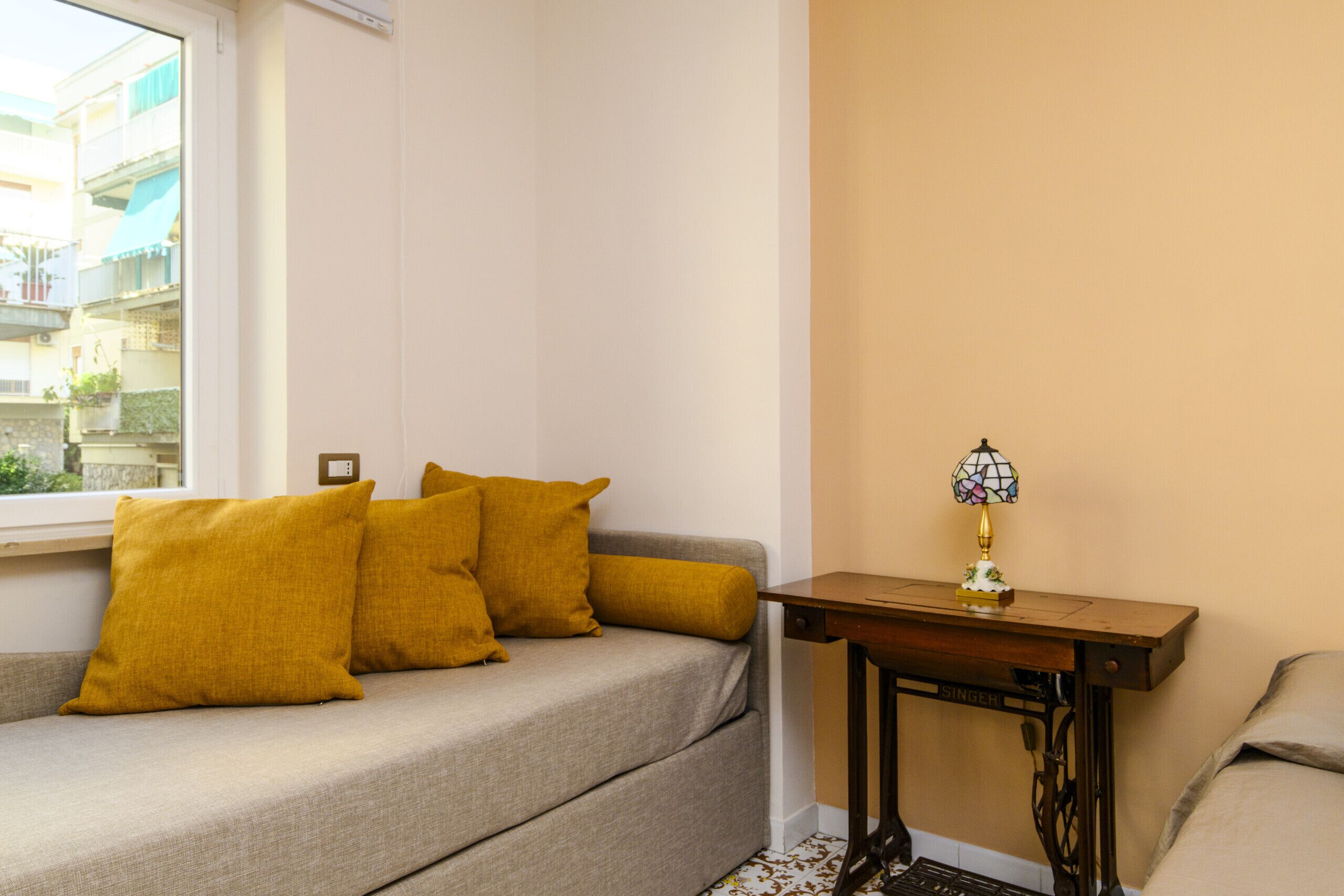 Una habitación con sofá, dos almohadas color mostaza, una mesita con una lámpara de vitral y una ventana con vistas a edificios.