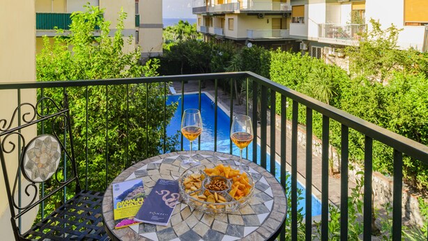Un balcón con vista a la piscina, una mesa con aperitivos y bebidas, y una revista.