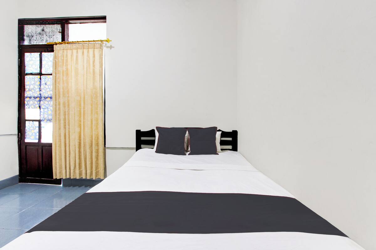 Ein Einzelbett mit schwarz-weißlicher Bettdecke, einem Holz-Kopfteil und einem Fenster mit Vorhang.