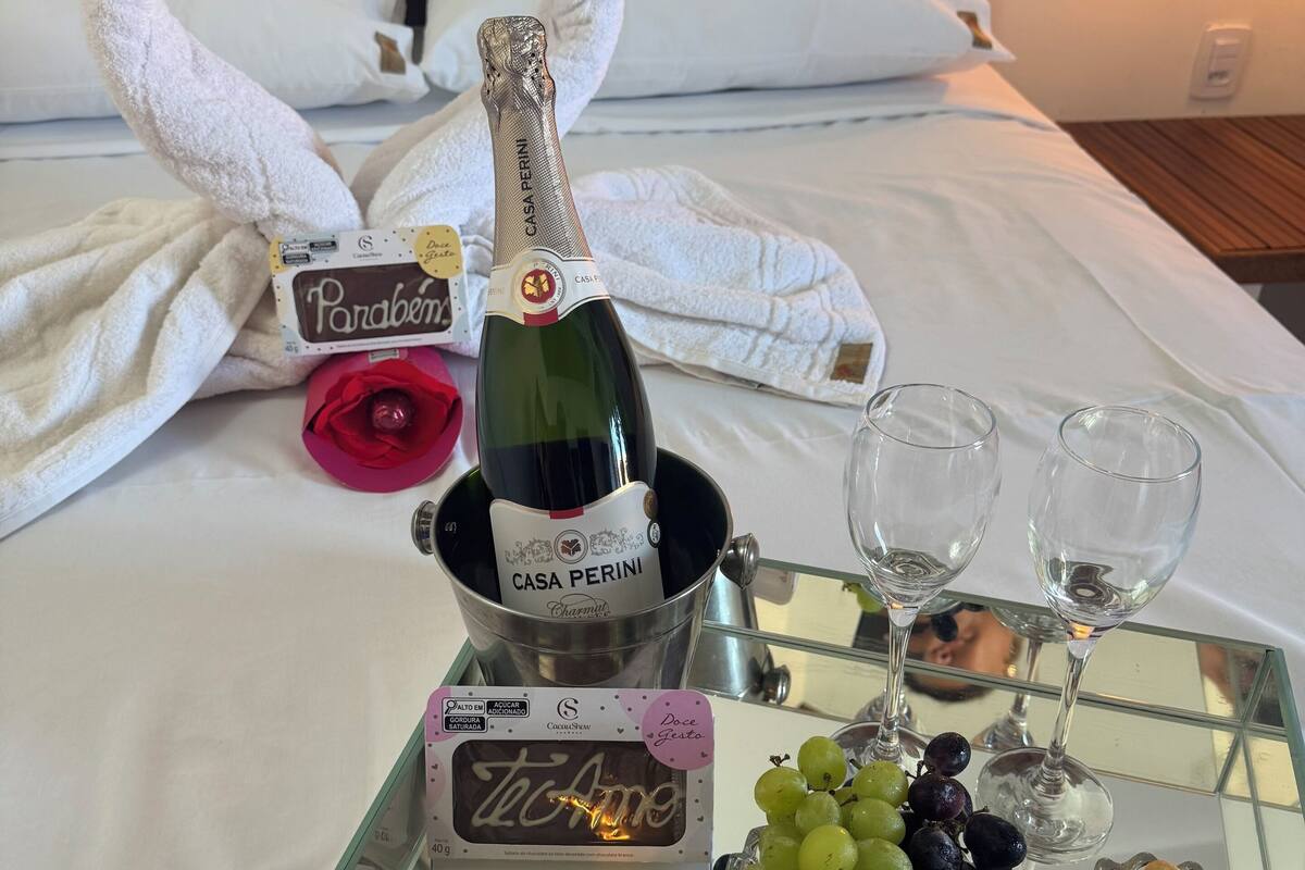 Un lit avec une bouteille de champagne Casa Perini, deux verres, un plateau de raisins et une barre de chocolat emballée.