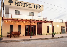 Un hotel llamado "HOTEL IPÉ" con un espacio de estar al aire libre techado, plantas en macetas y una fachada sencilla.
