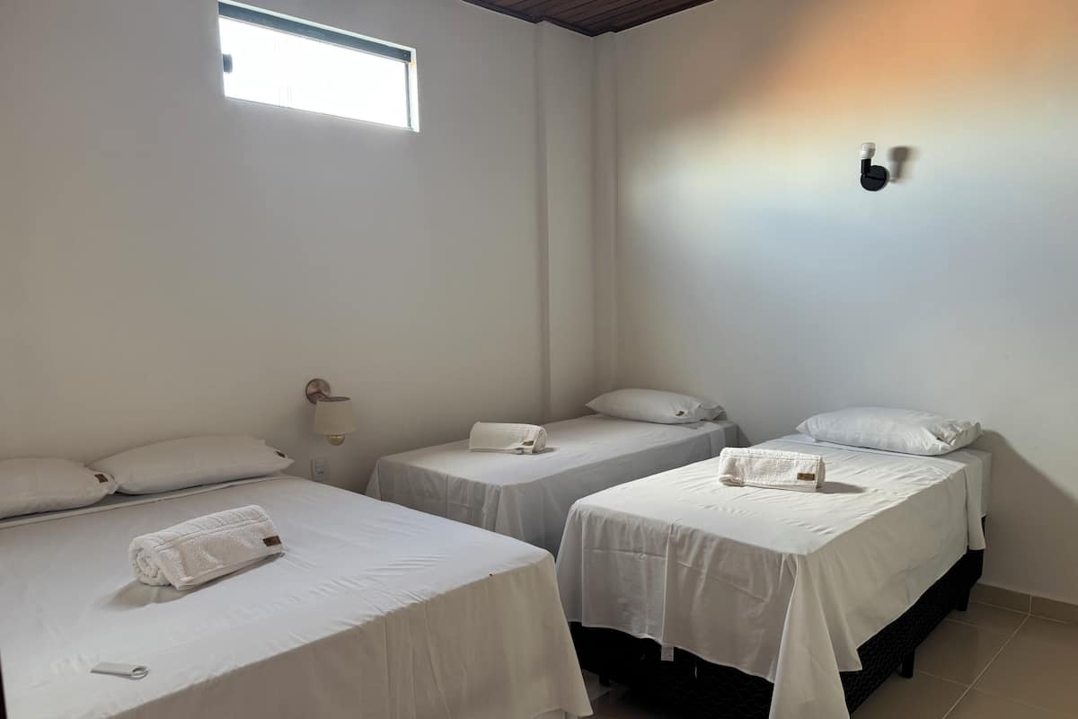 Une chambre avec trois lits simples, chacun recouvert d’une courtepointe blanche et agrémenté d’oreillers. Une lampe est fixée au mur sur le côté gauche, et une petite fenêtre se trouve au-dessus du lit situé à gauche.
