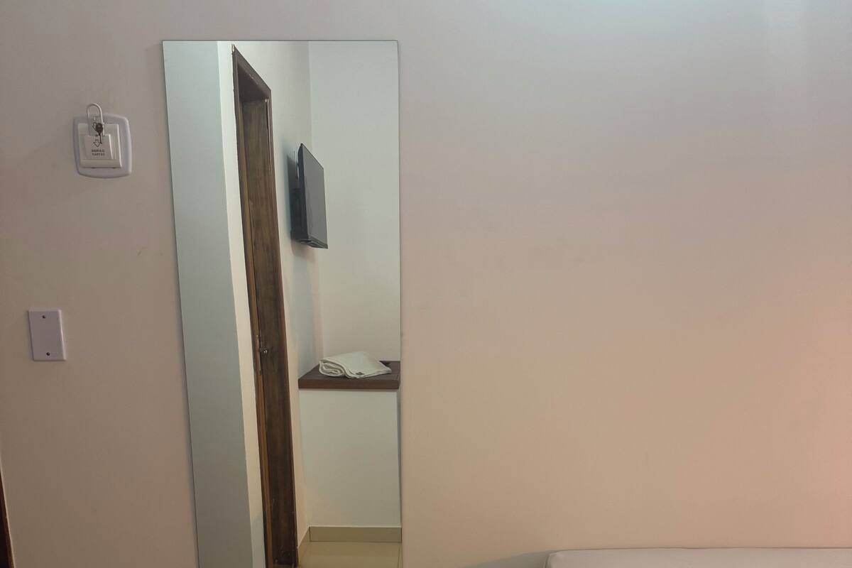 Une chambre à coucher avec un lit, un grand miroir, un téléviseur fixé au mur et une petite étagère en bois.