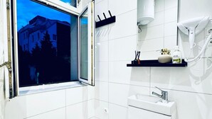 A bathroom with a window, a sink, a hairdryer, and a shelf with toiletries. - Compact Studio Apartment with Essentials, Ideal for 2 Guests (Wien)