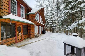A log cabin with a snow-covered exterior, a porch, and a view of a snowy forest. - Domaine Rivière Sacacomie - Le Huard (Saint-Alexis-des-Monts)
