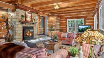 A log cabin interior with a stone fireplace, wooden beams, a checkered sofa set, and a television.