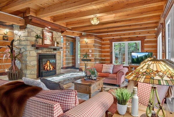 A log cabin interior with a stone fireplace, wooden beams, a checkered sofa set, and a television. - Domaine Rivière Sacacomie - Le Huard (Saint-Alexis-des-Monts)
